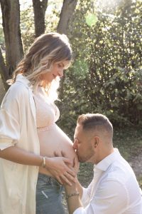 Couple attendant un bébé lors d'une séance photo grossesse en extérieur, le futur papa embrasse tendrement le ventre arrondi de la future maman dans une ambiance naturelle et lumineuse.